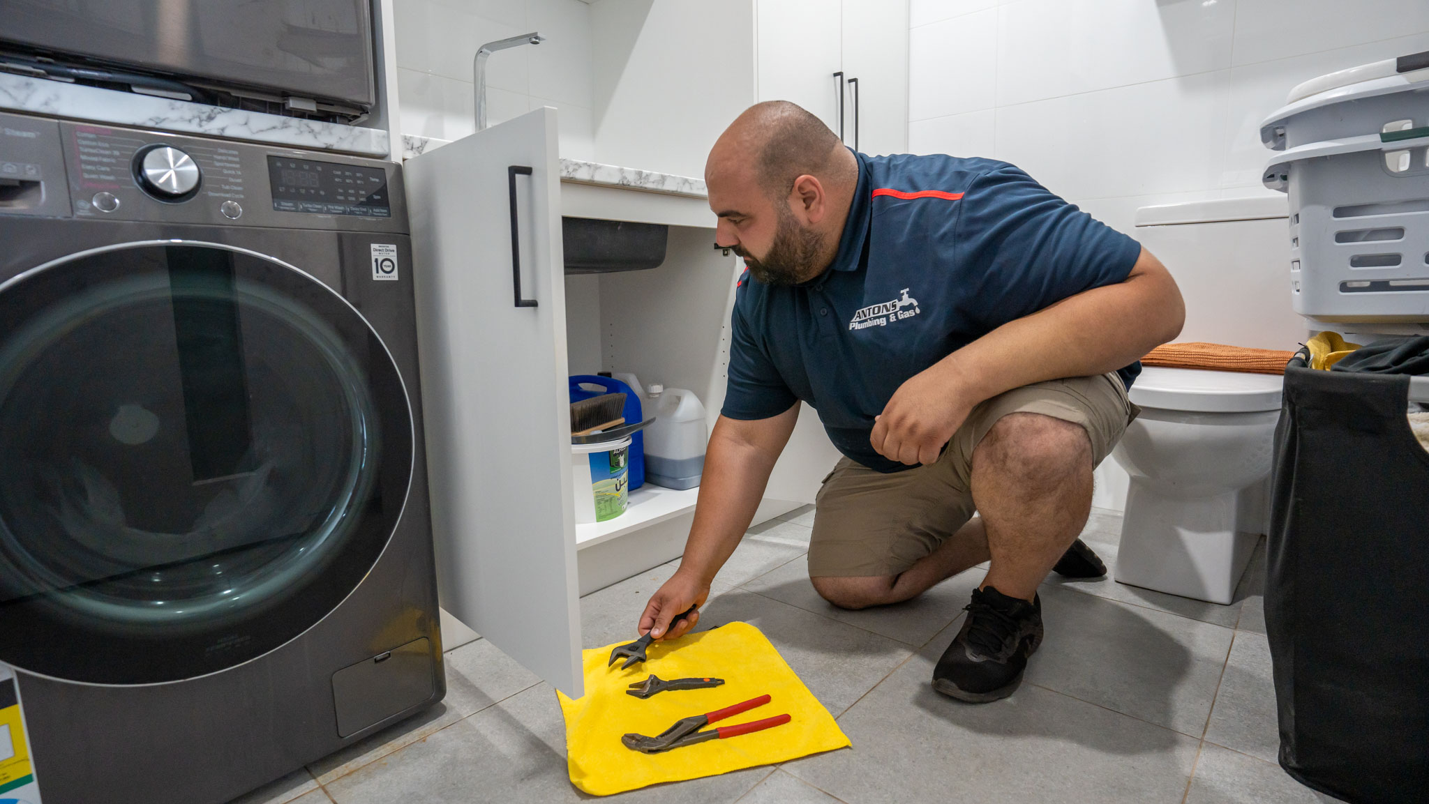A professional plumber from Anton's Plumbing examines under-sink plumbing in a laundry area, addressing potential burst pipes.